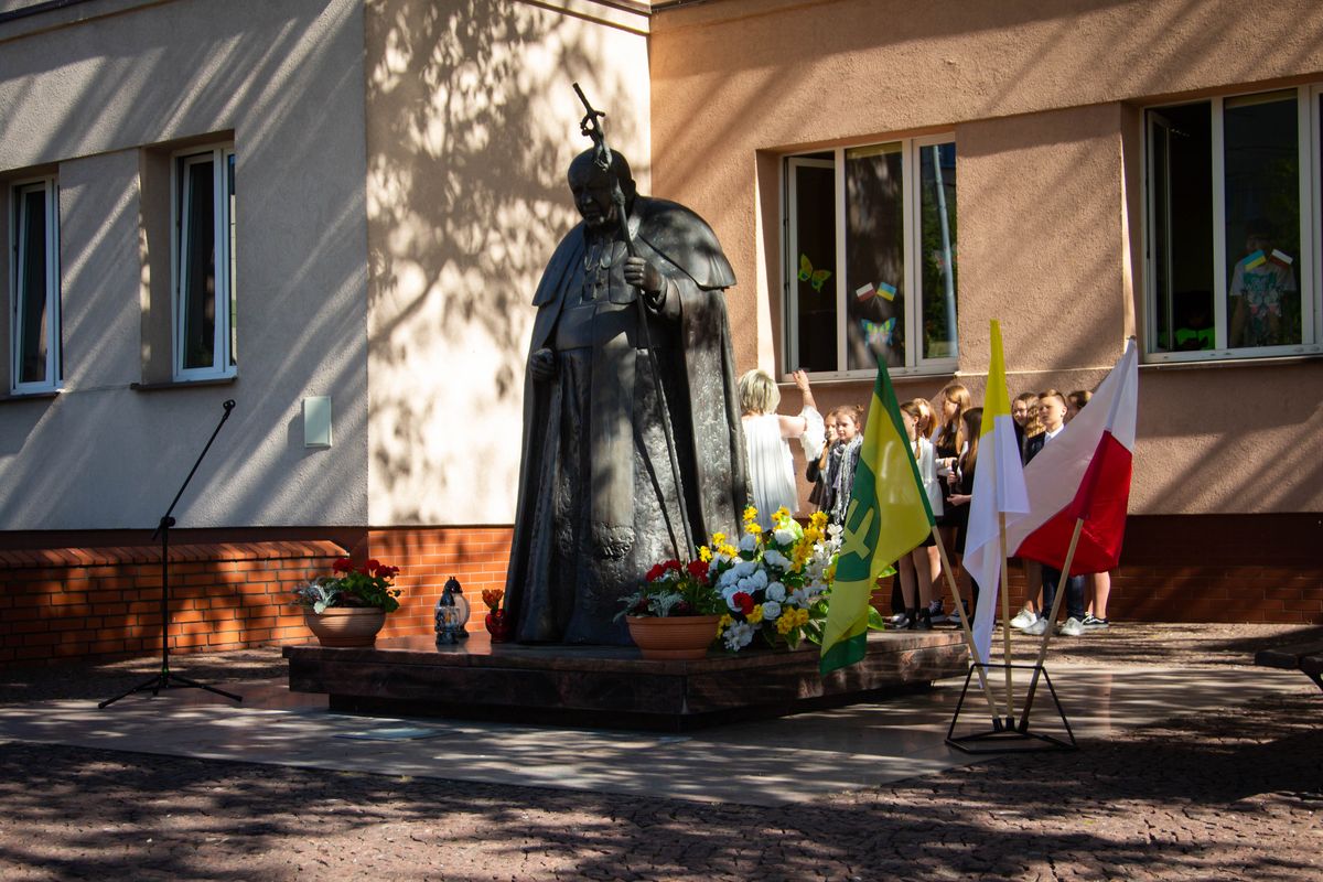 Pomnik św. Jana Pawła II oraz dzieci z panią nauczycielką śpiewają podczas święta szkoły