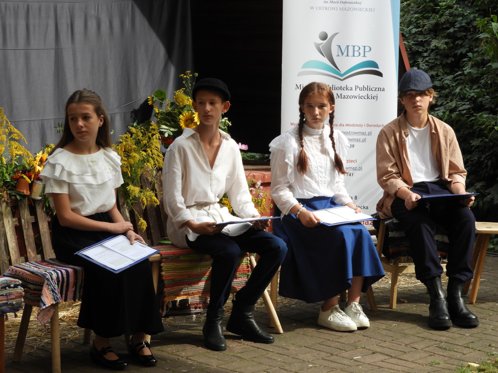 Czworo dzieci siedzących na przygotowanej scenerii czytających wybitne dzieła literatury polskiej