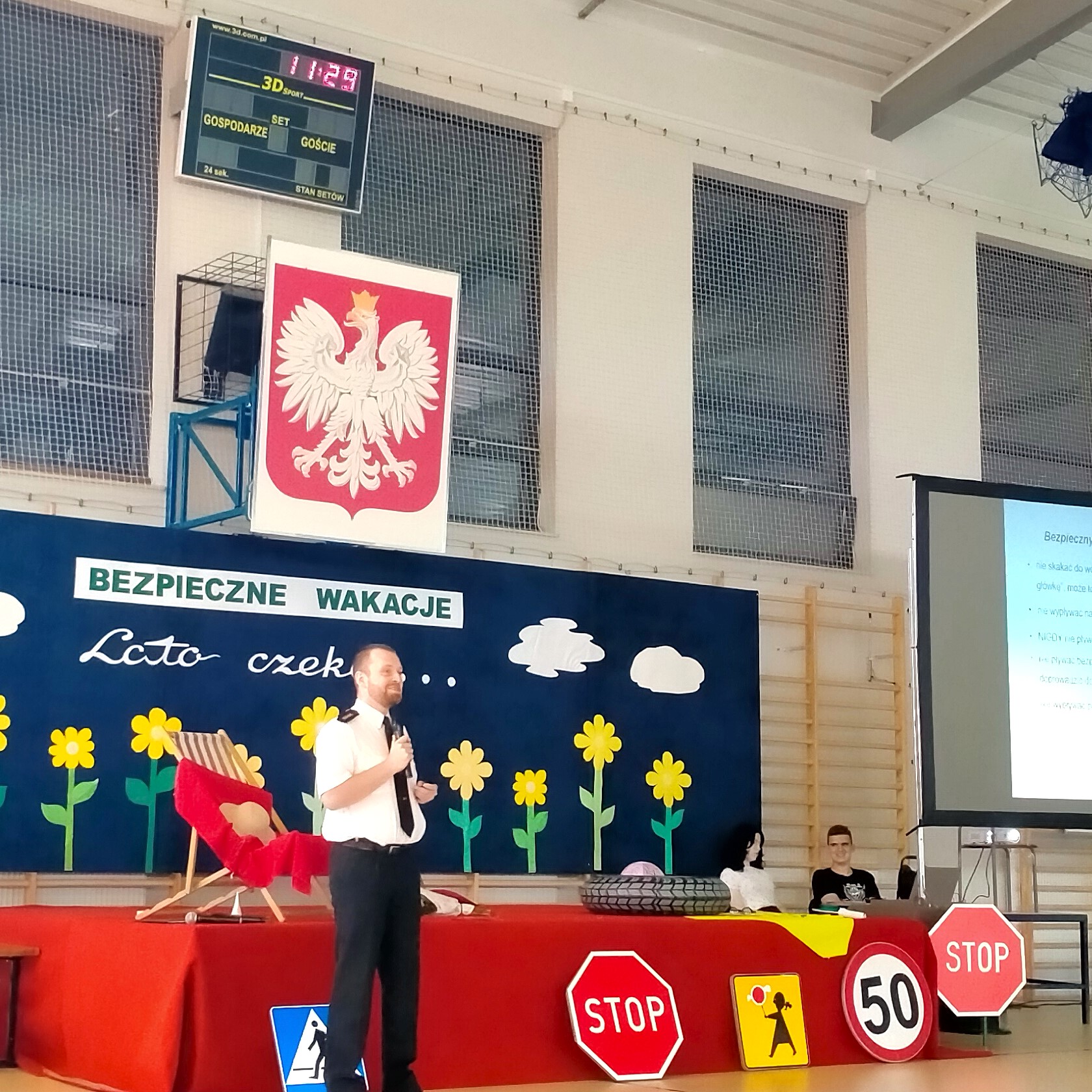 na zdjęciu policjant przemawia do mikrofonu na spotkaniu bezpieczne wakacje, które odbyło się w hali LO w Ostrowi Mazowieckiej, w tle napis bezpieczne wakacje, kwiaty, przed scena stoi scenografia dot. wakacji, znaki zakazu i nakazów