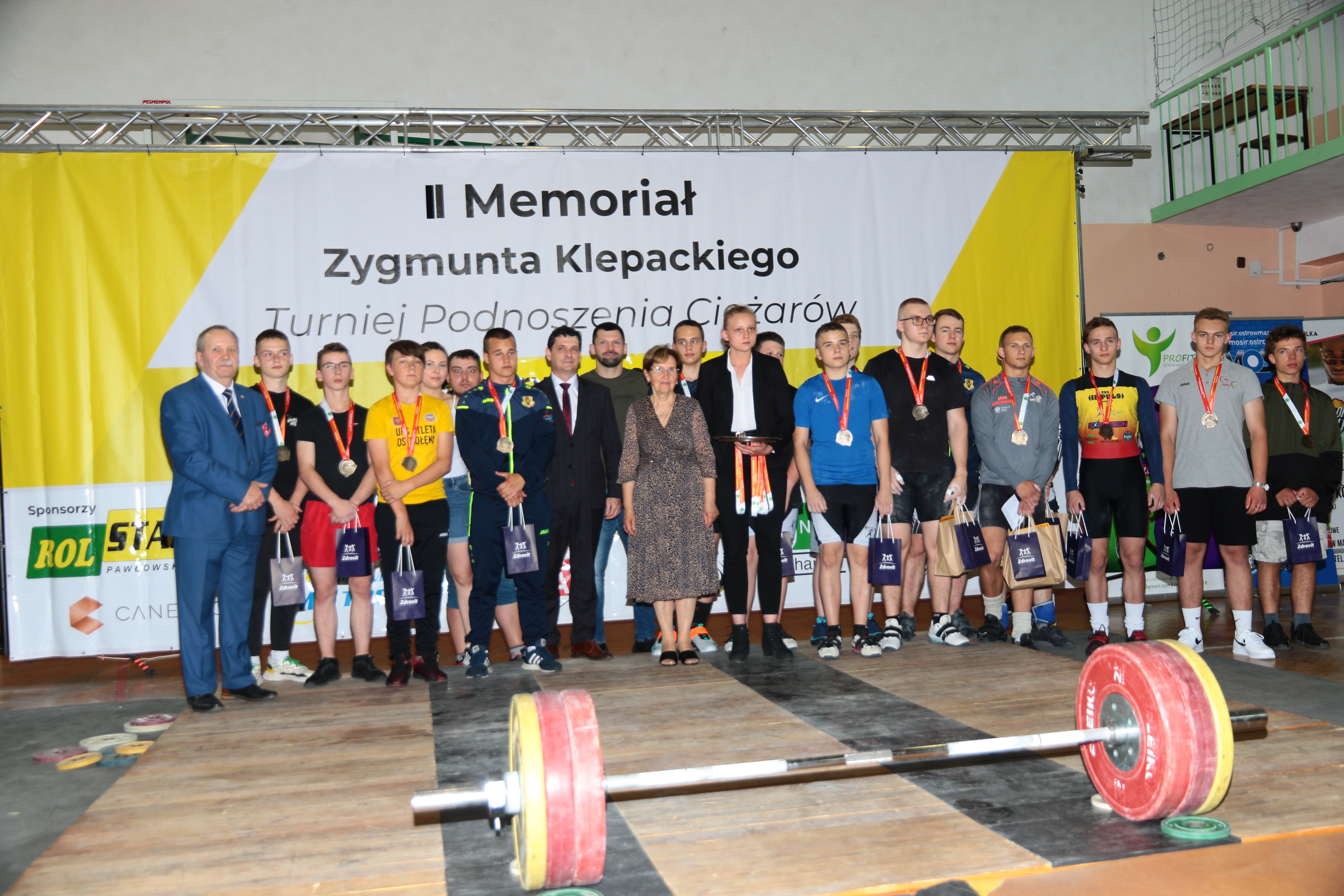 Grupa osób nagrodzonych na II Memoriale Zygmunta klepackiego, z medalami, dyplomami, sztanga lży na ziemi przed grupą