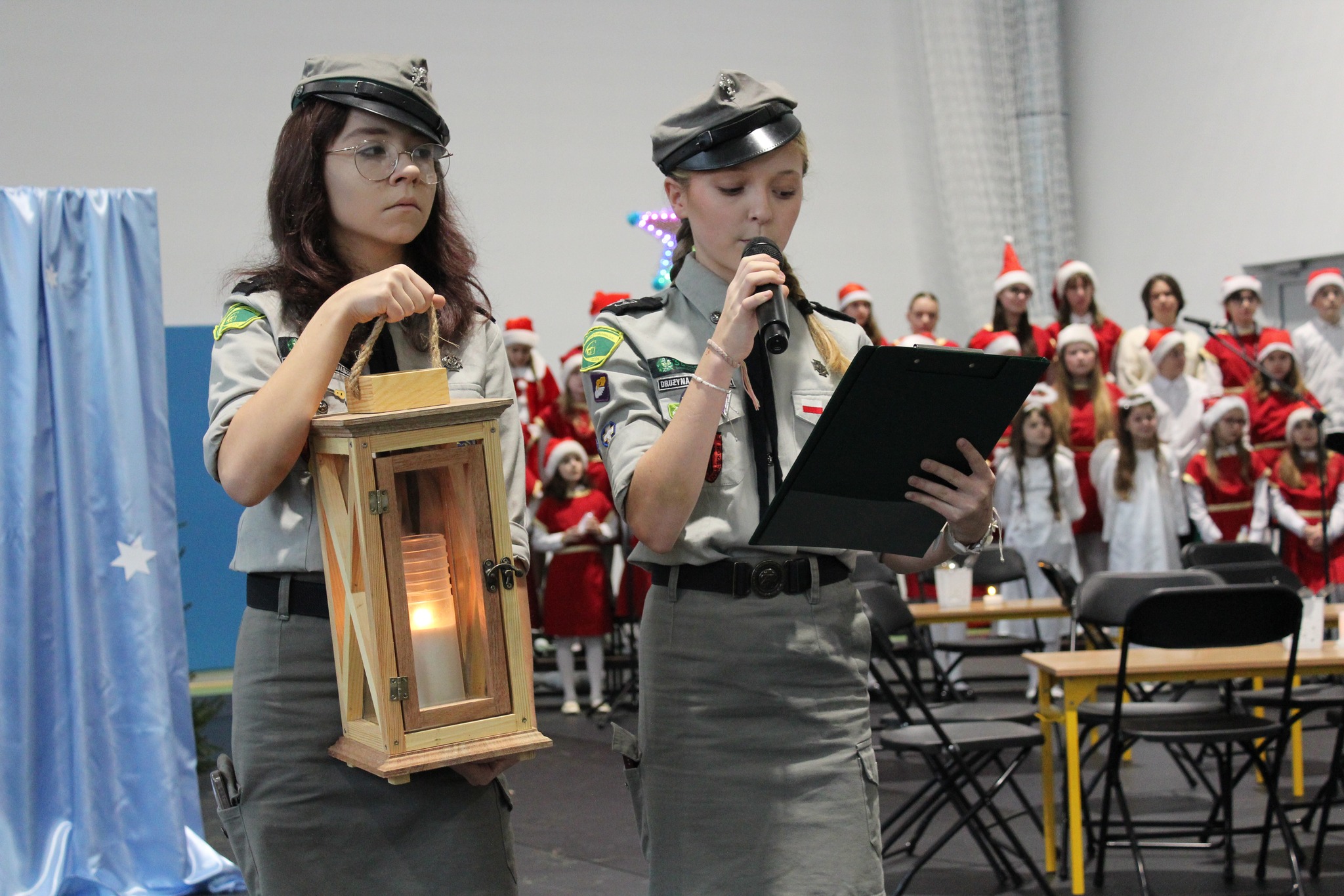 stoją dwie dziewczynki jedna z dziewczynek przemawia a druga trzyma betlejemskie światełko