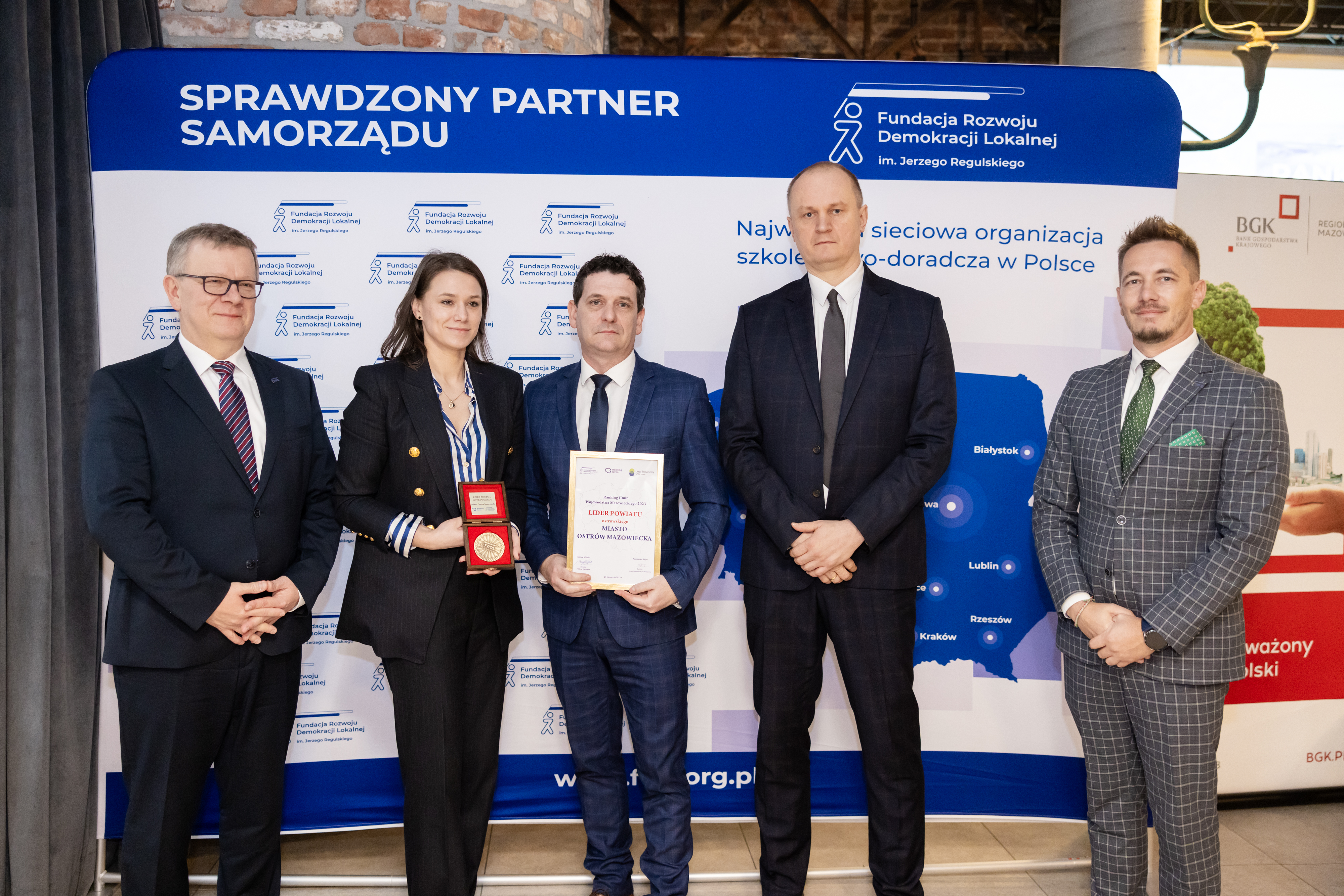 Dyrektor Wydziału Gospodarczego Małgorzata Rosińska i Zastępca Burmistrza Daniel Choinka trzymający nagrodę, wraz z gośćmi na tle ścianki promocyjnej Fundacji Rozwoju Demokracji Lokalnej