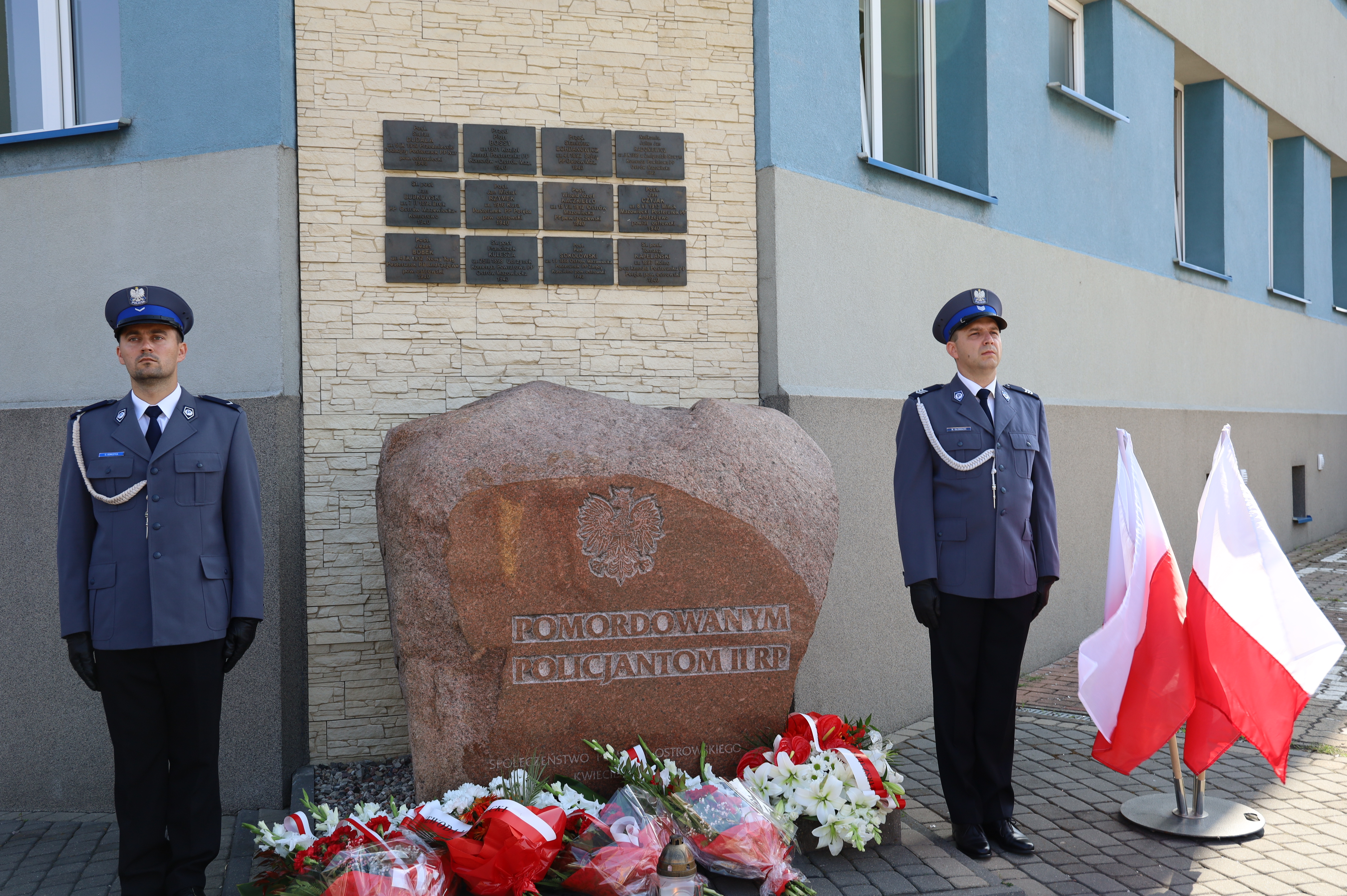 dwoch policjantów stoja przy pomniku pomordowanym policjantom II RP, złożone kwiaty pod pomnikiem, flagi państwowe stoją obok 